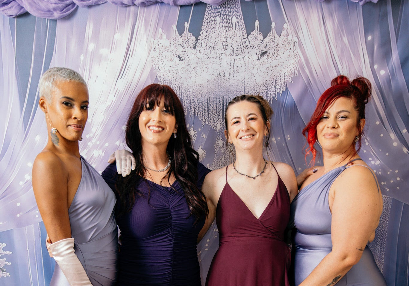 Four women posing together in elegant floor-length formal dresses. The group includes a woman with short platinum-blonde hair in a light lavender gown with white gloves, a woman with dark hair in a deep navy gown, a woman with light brown hair in a burgundy gown, and a woman with red hair in a light lavender gown.