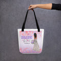 Black handle beige 15x15 canvas tote bag, with square image showing hearts in purple in pink shades, and Balloon text "Bride" in purple with "of Christ" in fancy text with "Proverbs 31" underneath and to the right is back side of Bride, medium skin tone, long dark hair, holding a purple bouquet.